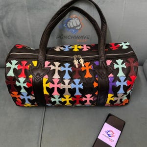 Peut inclure: Sac de voyage en cuir noir orné d'un motif de croix multicolores. Le sac est doté de deux poignées noires et d'une fermeture éclair. Un smartphone avec le texte "PUNCHWAVE" est à côté du sac.