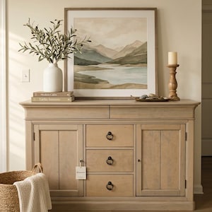 May include: A light brown wooden sideboard featuring two side cabinets and three central drawers with black ring pulls. A white vase with olive branches, two stacked books, a framed landscape print, and a wooden candle holder sit on top.