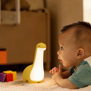 May include: A yellow dinosaur-shaped night light with a white glowing center. The lamp is on a soft surface with colorful toy blocks nearby. A baby is looking at the lamp.