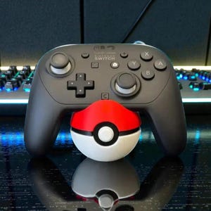 May include: A black Nintendo Switch Pro Controller with white joysticks and buttons, resting above a red, white, and black Pokeball. The controller has the Nintendo Switch logo.
