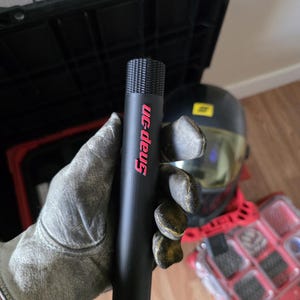 May include: A black Snap-on tool held in a gloved hand. The tool is cylindrical with a textured top and the Snap-on logo in red. A black toolbox, a welding helmet, and a red parts organiser are in the background.