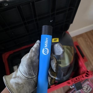 May include: A blue Miller welding rod holder with a black textured cap. The cylindrical holder is held by a gloved hand, with a black toolbox and welding helmet in the background.