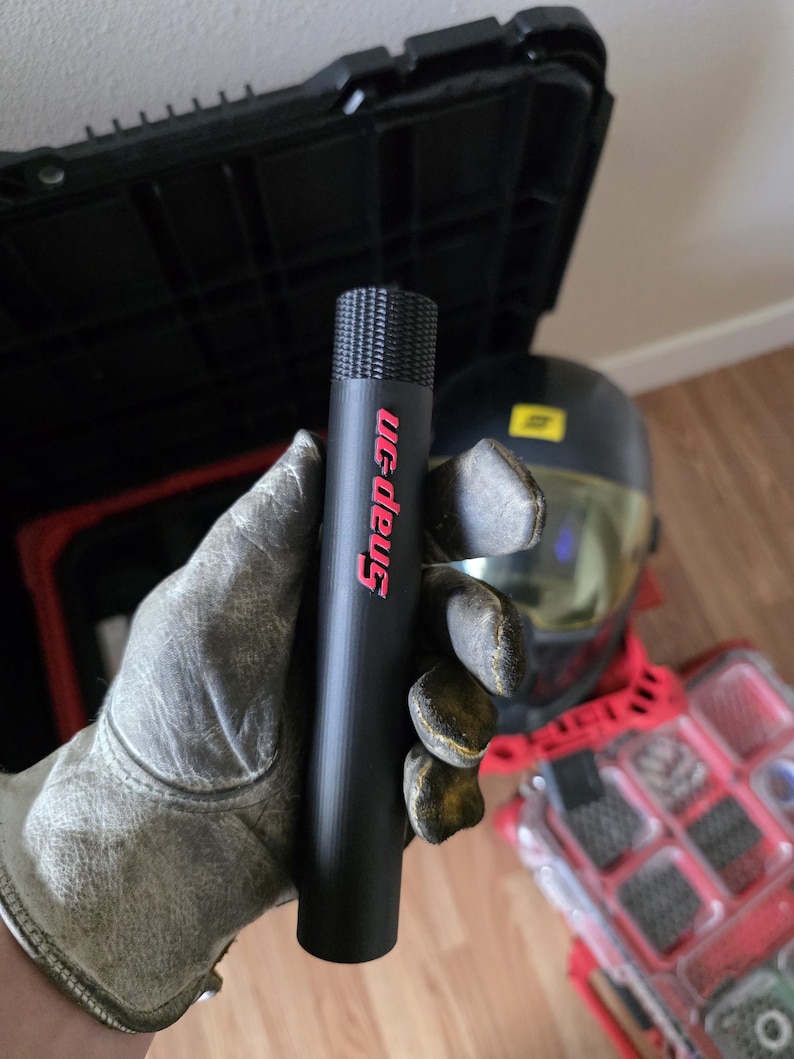 May include: A black Snap-on tool held in a gloved hand. The tool is cylindrical with a textured top and the Snap-on logo in red. A black toolbox, a welding helmet, and a red parts organiser are in the background.