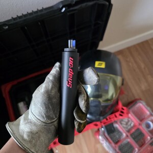 May include: A black Snap-on torch with red lettering is held by a gloved hand. The torch has a cylindrical shape. A black toolbox and a helmet are in the background, along with a red toolbox.