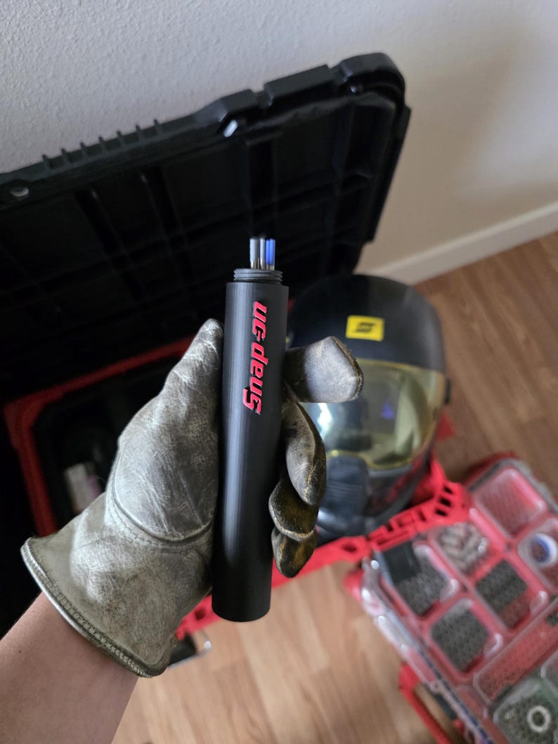 May include: A black Snap-on torch with red lettering is held by a gloved hand. The torch has a cylindrical shape. A black toolbox and a helmet are in the background, along with a red toolbox.