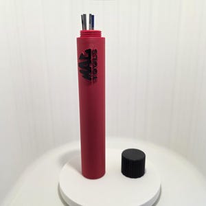 May include: A red cylindrical tool holder with the text "MAC TOOLS" in black. The holder contains several metal tools. A black cap sits next to the holder on a white circular base. The background is white.