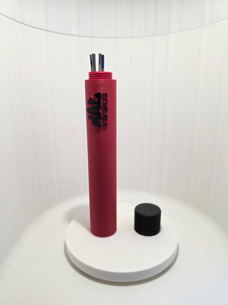 May include: A red cylindrical tool holder with the text "MAC TOOLS" in black. The holder contains several metal tools. A black cap sits next to the holder on a white circular base. The background is white.