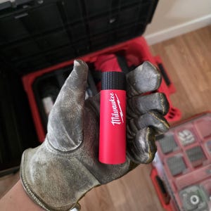 May include: A red and black cylindrical tool with the Milwaukee logo in white is held in a gloved hand. The tool has a textured black top and a red body. The background includes a red toolbox and a wooden floor.