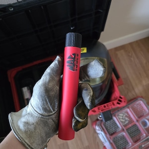 May include: A red cylindrical tool with the text "MAC TOOLS" in black. The tool has a textured black top. The tool is held by a person wearing a work glove. Other tools and a toolbox are in the background.