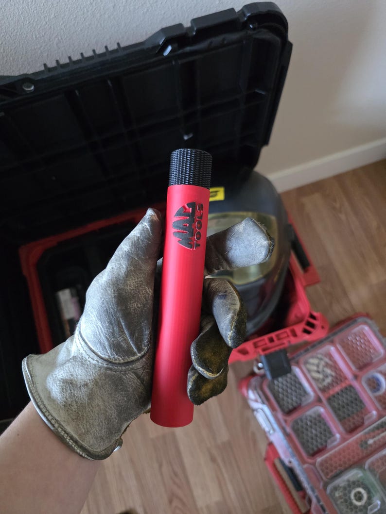 May include: A red cylindrical tool with the text "MAC TOOLS" in black. The tool has a textured black top. The tool is held by a person wearing a work glove. Other tools and a toolbox are in the background.