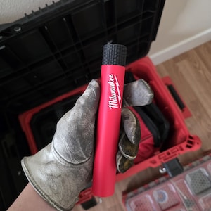 May include: A red cylindrical tool with the white "Milwaukee" logo. The tool has a textured black top and is held by a gloved hand. The background includes a black toolbox and a red toolbox.