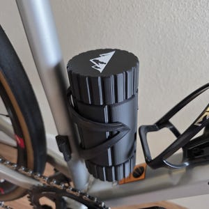 May include: Black cylindrical storage container with a mountain logo, mounted on a bicycle frame. The container is held in place by a black holder. The bicycle frame is silver.