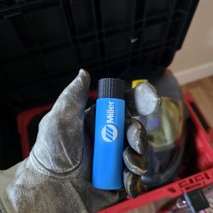 May include: A blue Miller welding torch with a black textured top. The torch is held in a work glove, with a welding helmet and toolbox in the background. The Miller logo is visible on the torch.