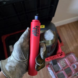 May include: A red cylindrical tool with the text "MAC TOOLS" in black. The tool has a blue tip and is held by a person wearing work gloves. A black toolbox and a red toolbox are visible in the background.