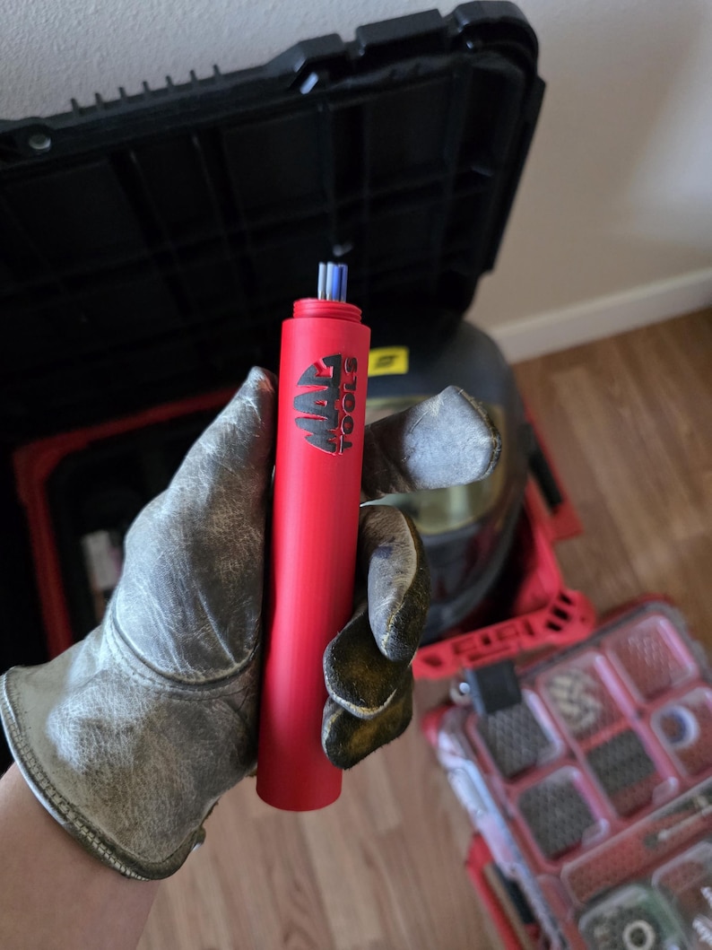 May include: A red cylindrical tool with the text "MAC TOOLS" in black. The tool has a blue tip and is held by a person wearing work gloves. A black toolbox and a red toolbox are visible in the background.