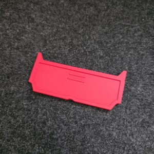 May include: A vibrant red rectangular item with a recessed central section and distinctive notched edges. The object, made from a smooth material, is set against a dark grey, textured backdrop.