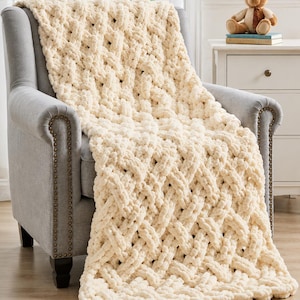 May include: A chunky, cream-colored knit throw blanket featuring a thick, braided cable pattern. The blanket is draped over a gray upholstered armchair with nailhead trim, showcasing a soft, textured yarn construction with large, interlocking loops.