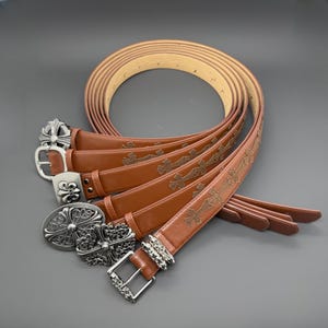 Op de afbeelding: Verschillende bruine leren riemen met decoratieve zilveren gespen worden getoond. De riemen hebben reliëf kruispatronen en verschillende gespontwerpen, waaronder sierlijke bloemen- en kruismotieven. De riemen zijn in een cirkelvorm gerangschikt.