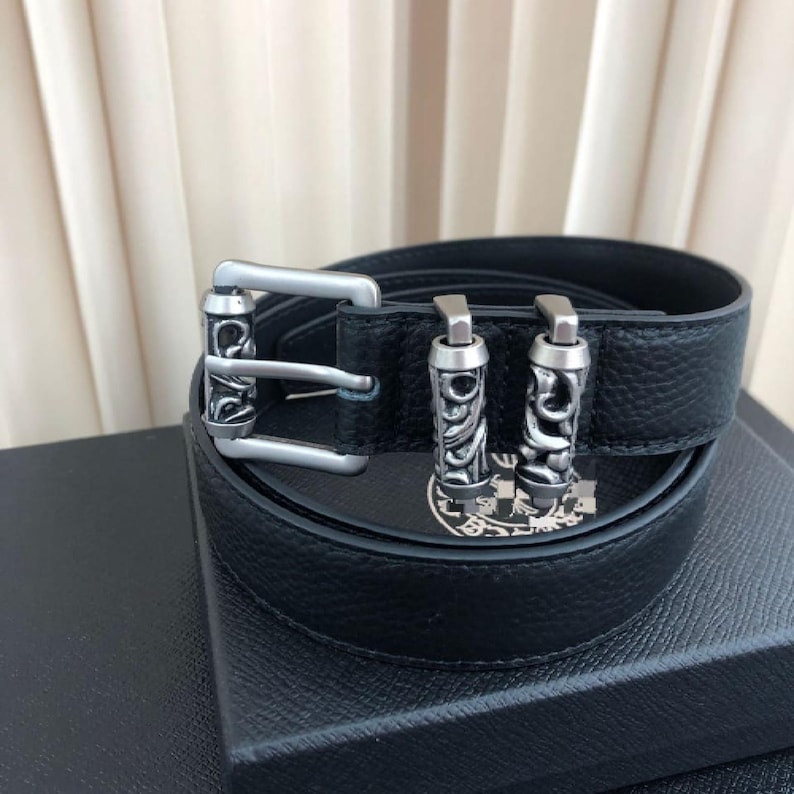 Pode incluir: Cinto de couro preto com fivela prateada e duas presilhas a condizer. A fivela tem uma moldura retangular com volutas decorativas. O cinto est&aacute; numa caixa preta.