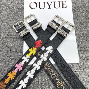 Peut inclure: Quatre ceintures en cuir noir avec des boucles argentées et des motifs de croix de différentes couleurs. Les ceintures sont disposées sur une surface blanche avec le mot "OUYUE" imprimé. Les motifs de croix comprennent le rouge, le jaune, le blanc et le marron.
