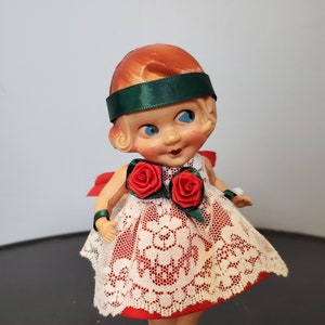May include: A vintage doll with red hair, wearing a red and white lace dress with green ribbon accents and red roses. The doll is wearing green shoes with white pom-poms.