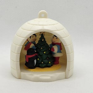 Puede incluir: Adorno navideño en forma de iglú color crema. En el interior, dos figuras con atuendo de invierno se encuentran a ambos lados de un árbol de Navidad decorado. También hay un pingüino con una bufanda roja. Una estrella dorada corona el árbol.