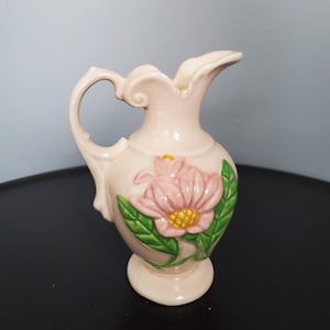 Può includere: Un piccolo brocca in ceramica rosa pallido con manico e beccuccio. La brocca è decorata con un fiore rosa in rilievo con centro giallo e foglie verdi. La brocca ha una base a piedistallo.