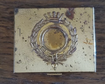May include: A vintage gold-toned metal compact with a raised design of a crown, wreath, and a circular center. The compact has a worn, aged appearance.