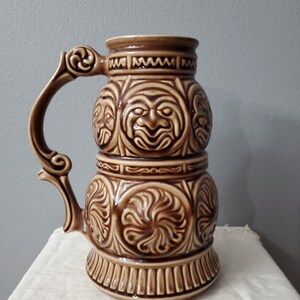 Pode incluir: Uma caneca de cerveja de cerâmica marrom com alça e entalhes intrincados de rostos e desenhos florais.