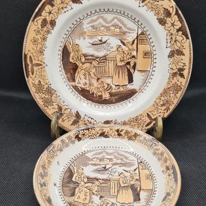 May include: Two brown and white ceramic plates with a floral border and a scene of people in a village setting. The plates are stacked on top of each other.