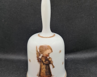 May include: White ceramic bell with a long handle and a gold-colored rim. The bell features a detailed illustration of an angel playing a flute, with gold stars scattered around the image. A decorative item.