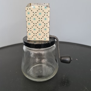 Könnte beinhalten: Ein vintage Gewürzmühle aus Glas mit schwarzem Deckel und Handkurbel. Die Mühle hat ein weißes Metall-Oberteil mit einem blauen und weißen geometrischen Muster.