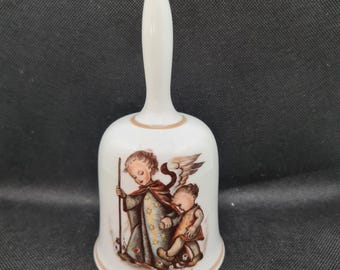 May include: A white porcelain bell with a brown handle and a painted image of an angel with wings guiding a child. The image is in a vintage style with brown, yellow, and blue colors.