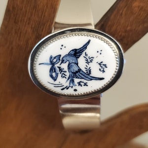 Op de afbeelding: Zilverkleurige manchetarmband met een ovale, witte keramische middenstuk. Het keramiek heeft een blauw vogelontwerp met bloemaccenten. De armband wordt gepresenteerd op een houten oppervlak.