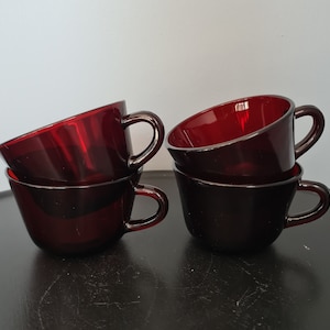 May include: Four ruby red glass teacups stacked in pairs. Each cup has a rounded bowl shape with a curved handle. The cups are on a dark surface, with a light gray background.
