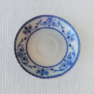 Puede incluir: Un platillo blanco con un diseño floral y de volutas azul alrededor del borde y un círculo central. El patrón azul incluye flores estilizadas y líneas curvas. El platillo probablemente está hecho de cerámica o porcelana.