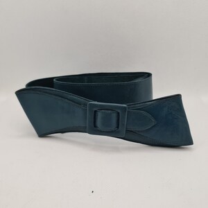 May include: A teal blue leather belt with a large, rectangular buckle. The belt is designed with a bow shape.
