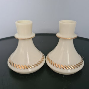 Puede incluir: Dos candelabros de cerámica blanca con ribetes dorados. Los candelabros tienen una base acampanada y una parte superior cilíndrica.