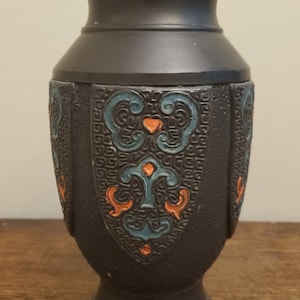 Puede incluir: Un jarrón de cerámica negro con un diseño geométrico en tonos azules y naranjas. El jarrón tiene una base ancha y un cuello estrecho.