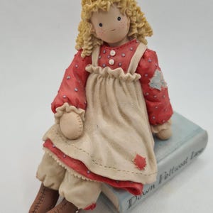 May include: A figurine of a girl with curly blonde hair, wearing a red dress with white polka dots and a white apron. She is sitting on a blue book with the title "Down Petticoat Lane". The figurine has brown boots and a sweet expression.