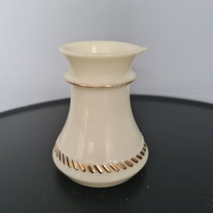 Puede incluir: Un pequeño jarro de cerámica de color crema con un borde dorado alrededor de la parte superior e inferior. El jarro tiene un labio ligeramente acampanado y un cuerpo redondeado.