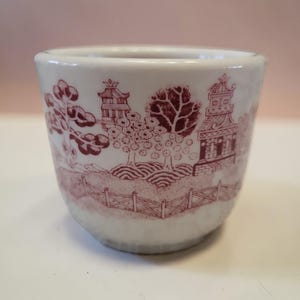 May include: A white ceramic egg cup with a maroon landscape design featuring trees, pagodas, and a fence. The cup has a smooth, glossy finish and a slightly flared shape. The design is a classic, vintage style.