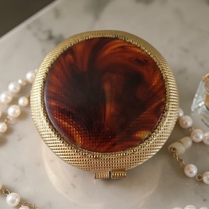 May include: A vintage gold-toned compact with a tortoiseshell design on the lid. The compact is round with a textured border. A strand of pearls and a perfume bottle are also in the image.