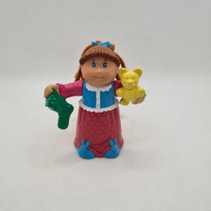 Può includere: Una bambola Cabbage Patch Kid con un vestito a pois rosa e bianco, scarpe blu e un maglione blu e bianco. La bambola tiene una calza verde e un orsacchiotto di peluche giallo.
