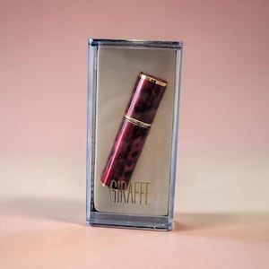 Puede incluir: Un tubo de lápiz labial granate y dorado en una funda de plástico transparente. El lápiz labial tiene un patrón veteado y detalles dorados. La funda tiene la palabra "GIRAFFE" impresa.