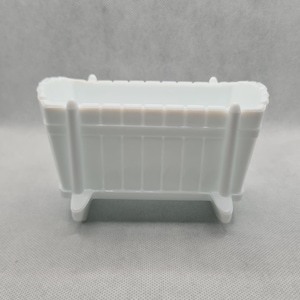 May include: A white plastic crib with a slatted design and four legs. The crib is a miniature version of a traditional crib.