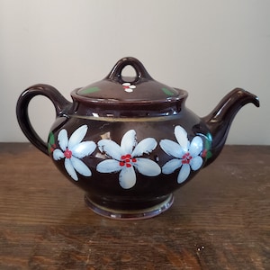 Puede incluir: Una tetera de cerámica marrón con tapa, asa y boquilla. La tetera está decorada con flores blancas con centros rojos. Acentos verdes están en la tapa y cerca del asa. La tetera está sobre una superficie de madera.