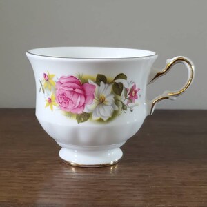 May include: White teacup with a pink rose and floral design. The cup has a gold handle and trim. The teacup is sitting on a wooden surface. The teacup is a vintage style.