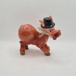 Könnte beinhalten: Eine Keramikfigur eines rosa Schweins, das einen schwarzen Zylinder trägt. Das Schwein hat einen weißen Bart und steht auf allen vieren.