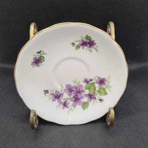 May include: A white porcelain saucer with a gold rim and a floral design featuring purple violets on a green stem.